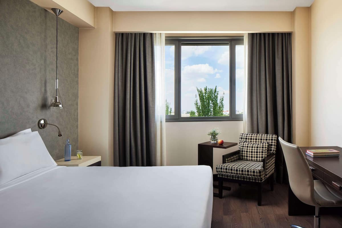A hotel room with a bed, a desk, a chair, a window with curtains, a small table with a bottle and glass, and a hanging light fixture.