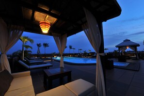 Zona de piscina al aire libre con sillones de descanso y un jacuzzi, rodeada de palmeras y un pabellón.