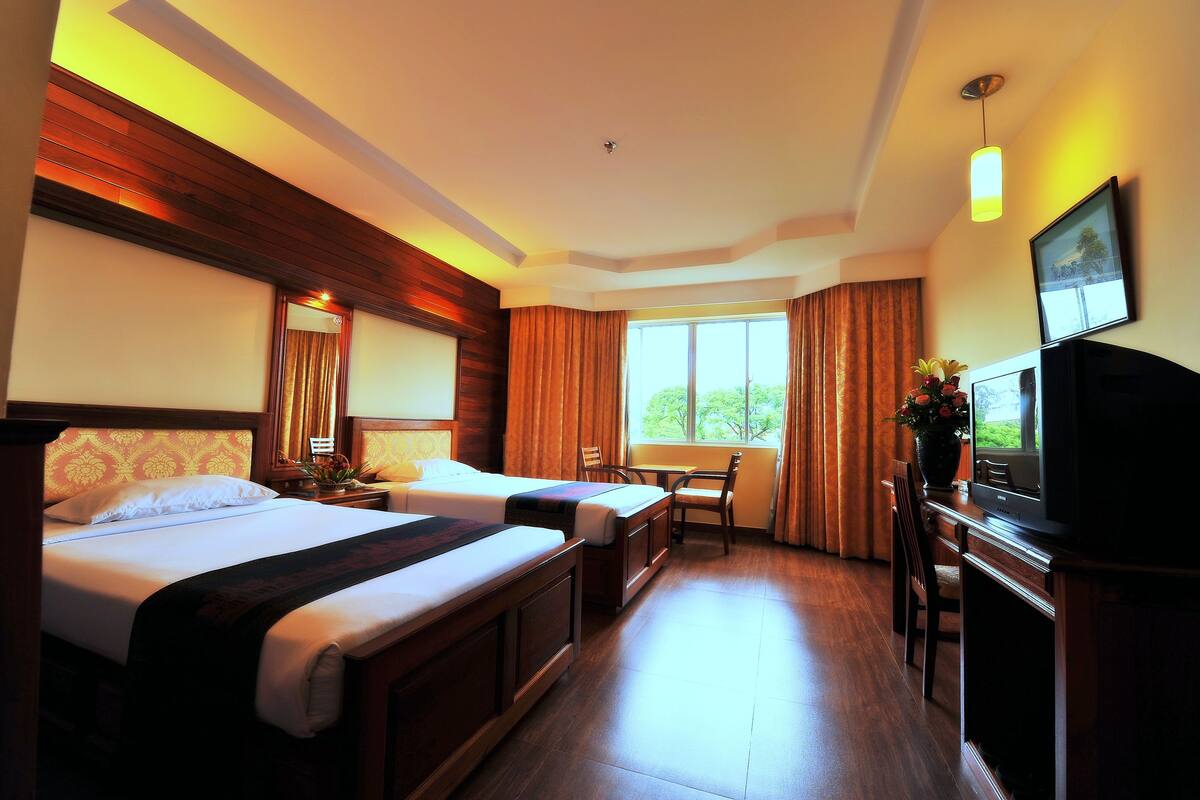 Quarto de hotel com duas camas, uma escrivaninha com televisão, uma janela com cortinas e vista para áreas verdes.