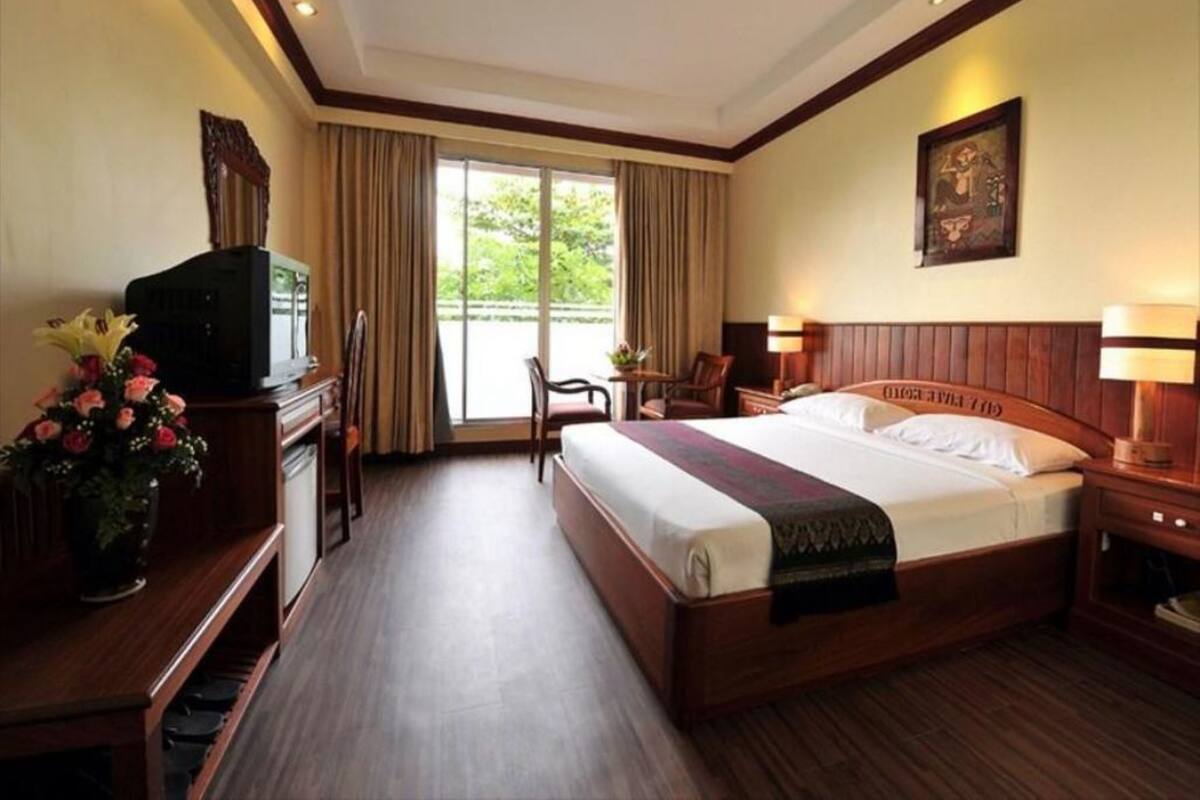 Quarto de hotel com uma cama grande, uma mesa com televisão, uma cadeira, abajur e uma janela com cortinas.