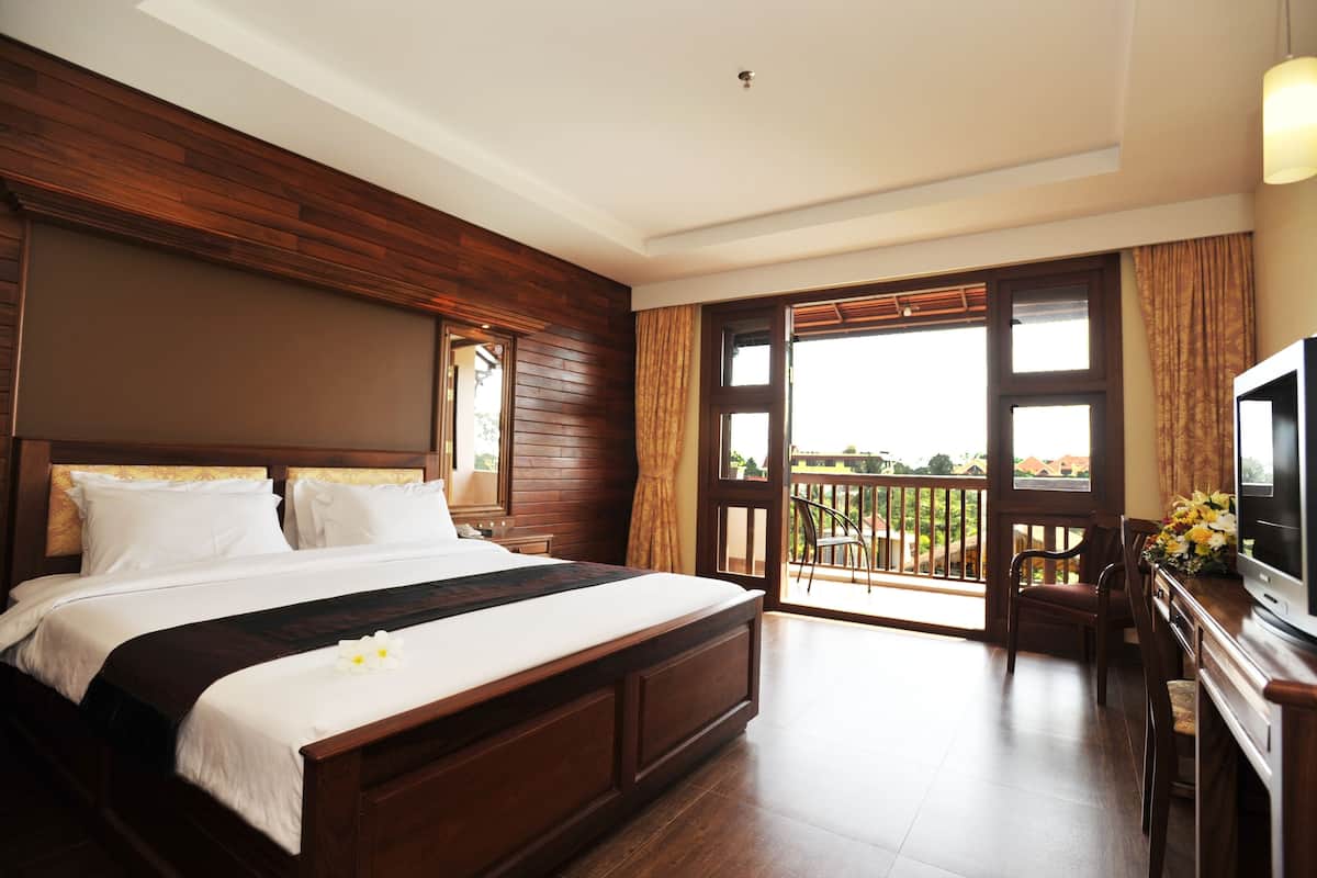 Quarto de hotel com uma cama grande, cabeceira de madeira, uma varanda com vista e uma televisão sobre um suporte de madeira.