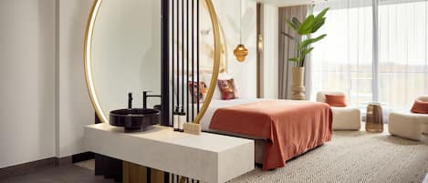 Una habitación de hotel moderna con un espejo grande, una mesita de noche con un cuenco negro, una cama con colcha naranja y una planta grande en maceta.