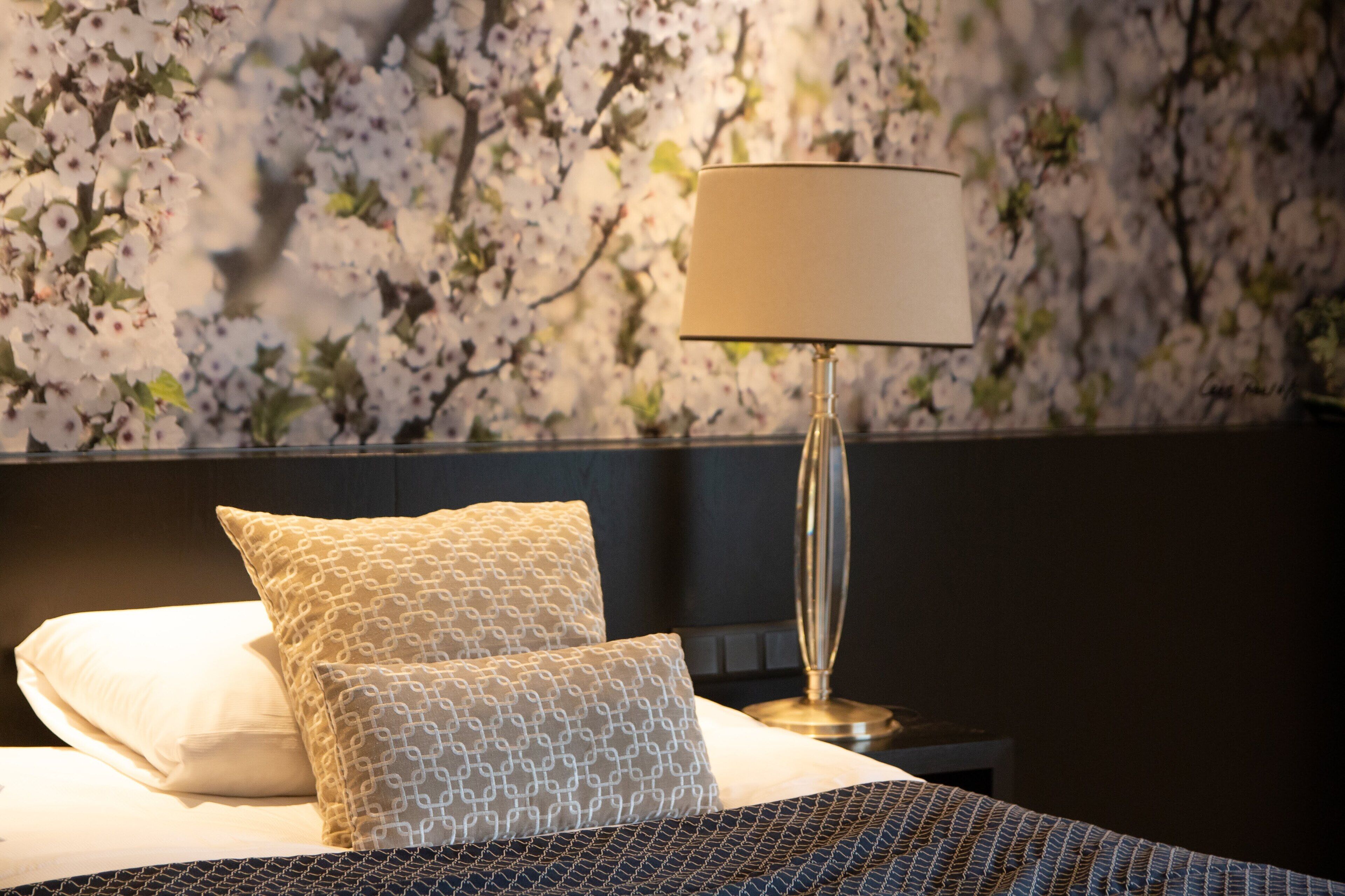 A neatly made bed with patterned pillows, a bedside lamp, and a floral wallpaper background.
