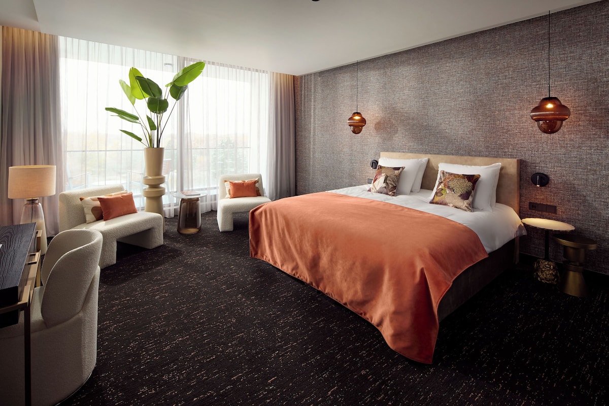 Ein modernes Hotelzimmer mit einem großen Bett, einer Sitzecke mit Sofa und Sesseln, einem Schreibtisch mit Lampe und einem großen Fenster mit Vorhängen.