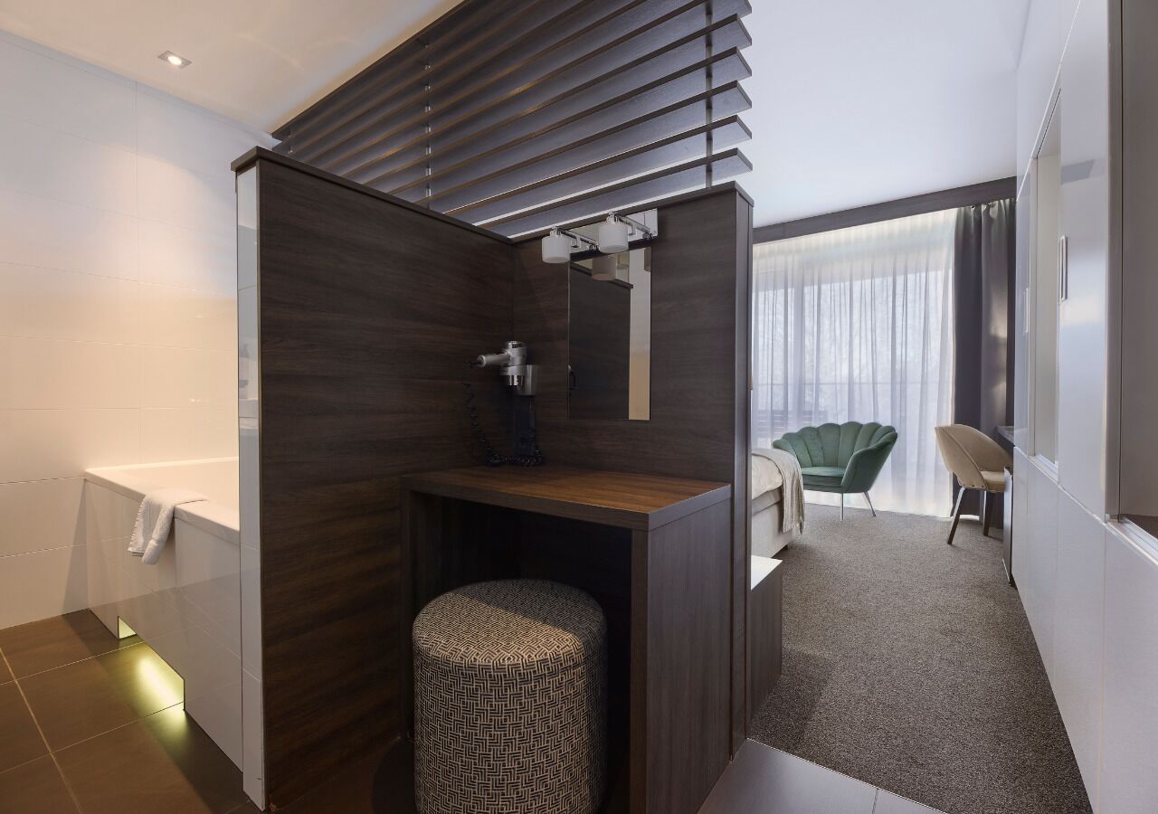 A modern bathroom with a shower, a toilet, and a vanity with a mirror. There is a chair and a small table in the adjacent room.