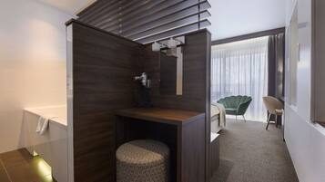 A modern bathroom with a shower, a toilet, and a vanity with a mirror. There is a chair and a small table in the adjacent room.