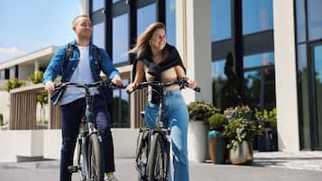 Deux personnes font du vélo sur une piste aménagée devant un bâtiment moderne doté de grandes fenêtres et de plantes en pot.