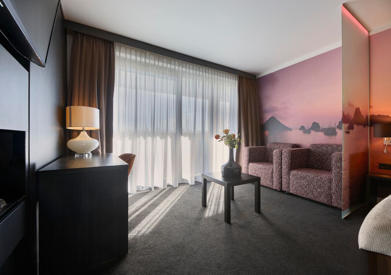 Quarto de hotel com televisão, uma escrivaninha com luminária, sofá, um vaso com flores e um mural na parede.