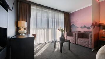 Quarto de hotel com televisão, uma escrivaninha com luminária, sofá, um vaso com flores e um mural na parede.