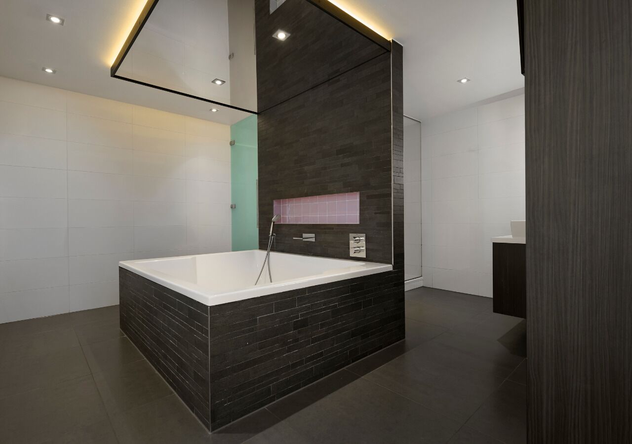 A modern bathroom with a large bathtub, dark tiled walls, and a mirrored ceiling.