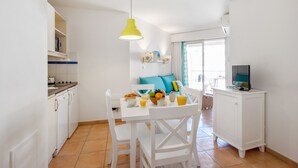 Una cocina luminosa con armarios blancos, una mesa de comedor con sillas, una lámpara colgante amarilla y un televisor sobre un mueble blanco.