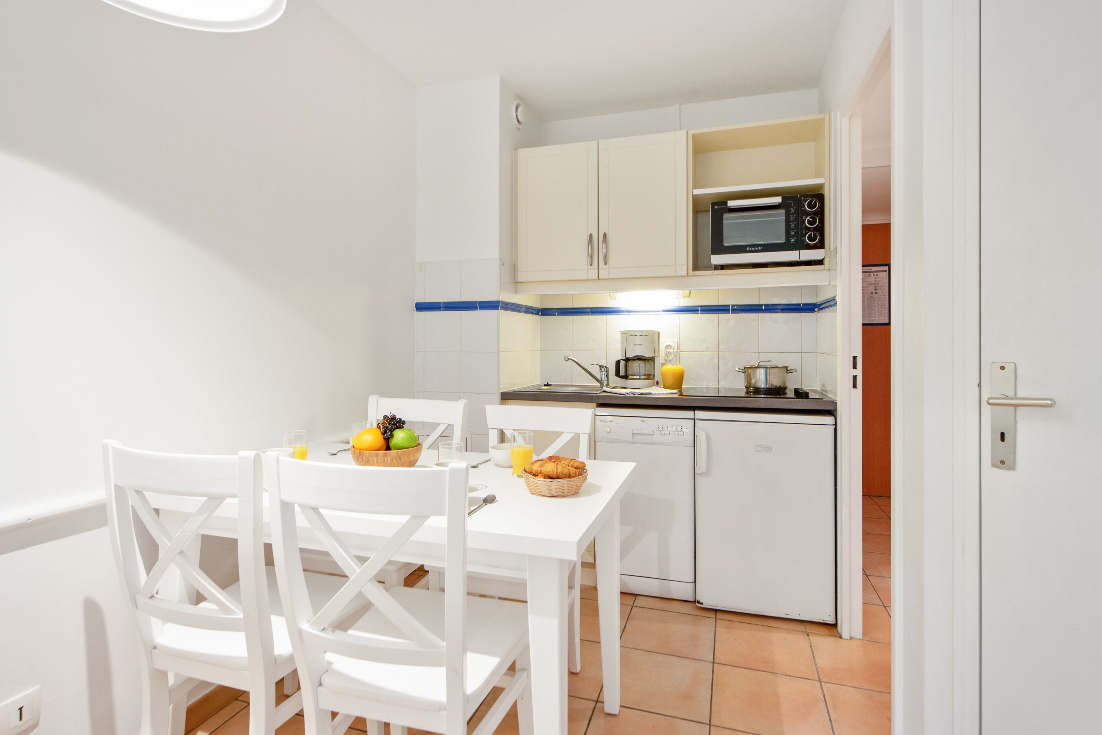 A small, modern kitchen with white cabinets, a microwave, and a coffee maker. There is a dining table with chairs and a basket of fruit.