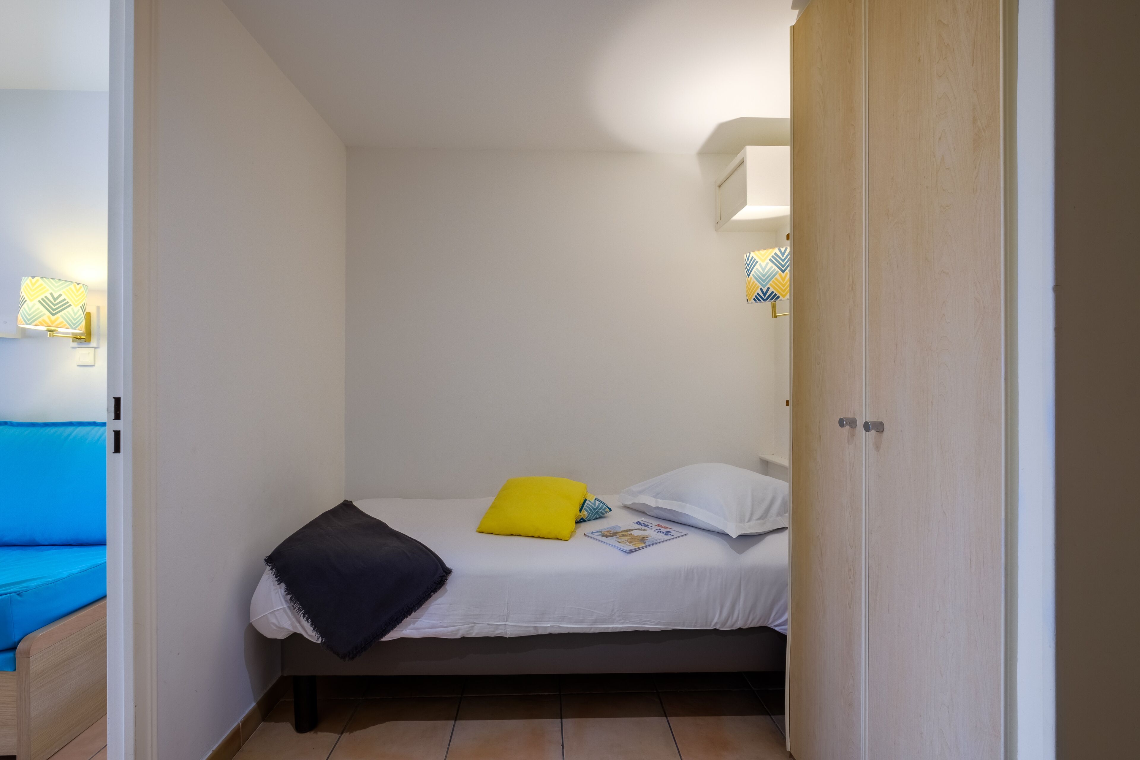 Une petite chambre avec un lit simple, recouverte d’une housse de couette blanche, un oreiller jaune et un plaid noir. Une porte en bois se trouve à droite, et un luminaire est fixé au-dessus du lit.