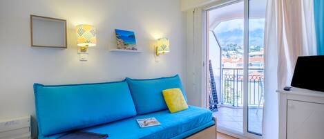 A bright room with a blue sofa, a TV, and a balcony with a view of buildings.