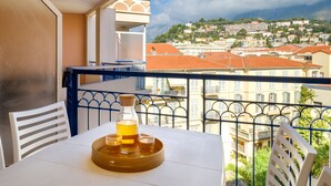 Un balcón con una mesa lista para dos personas, una botella de licor y dos vasos para chupitos. El balcón da a una ciudad con edificios de tejados rojos y un cielo despejado.