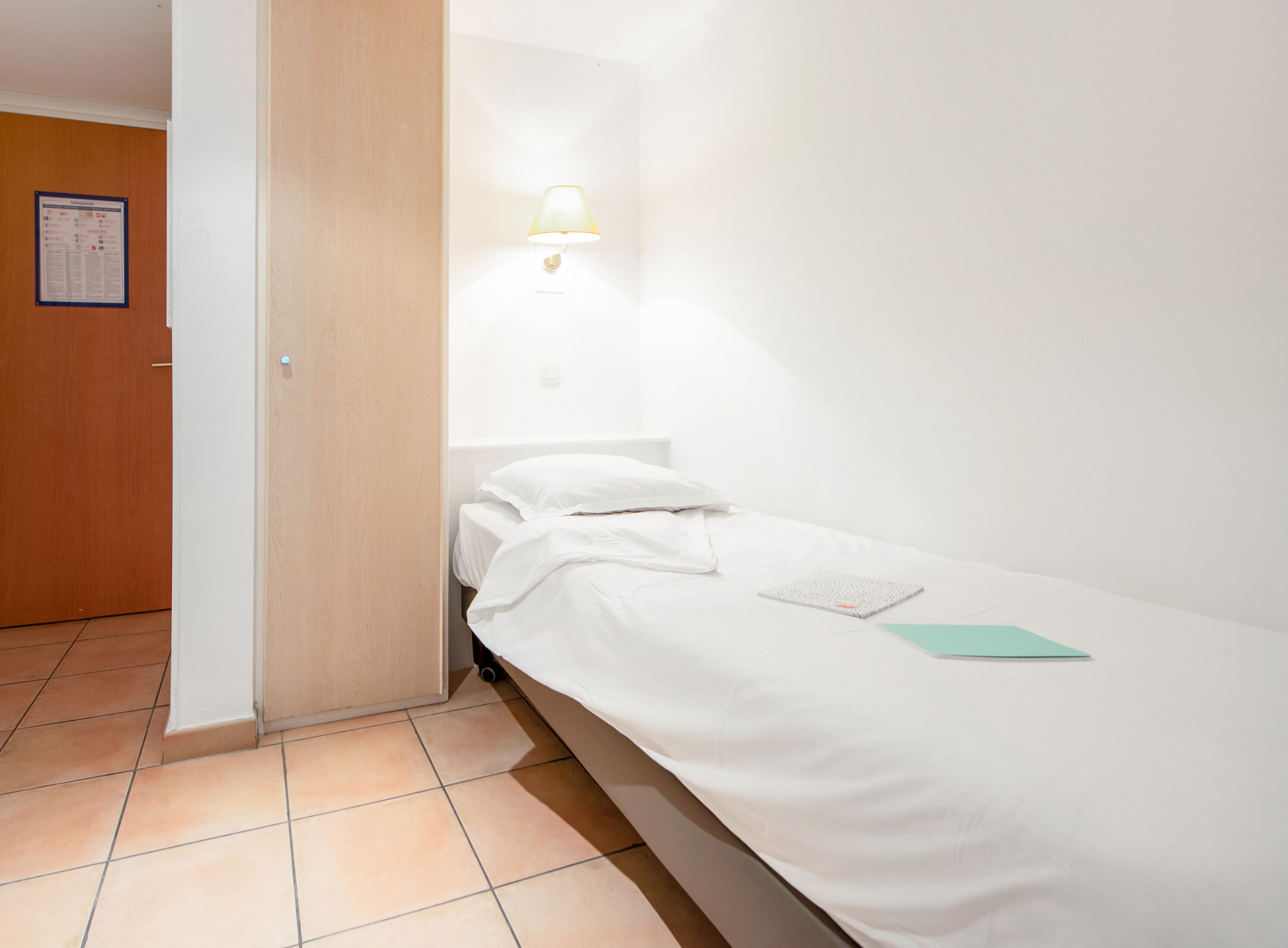 Un petit lit bien fait, avec des draps blancs et un seul oreiller, une petite table sur laquelle repose un livre et une lampe fixée au mur.