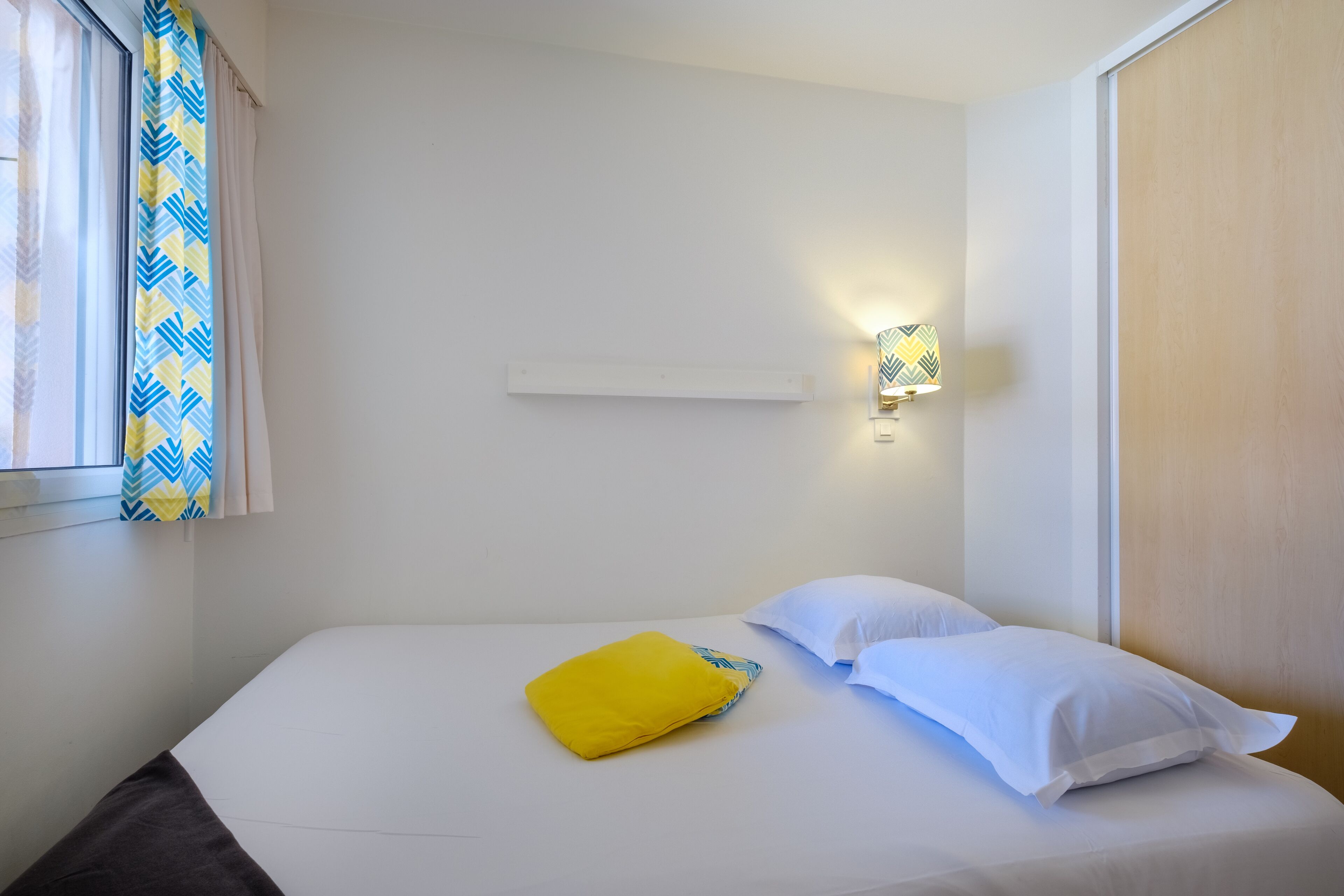 A single bed with white bedding and a yellow pillow, a bedside table, a wall-mounted light, and a window with curtains.