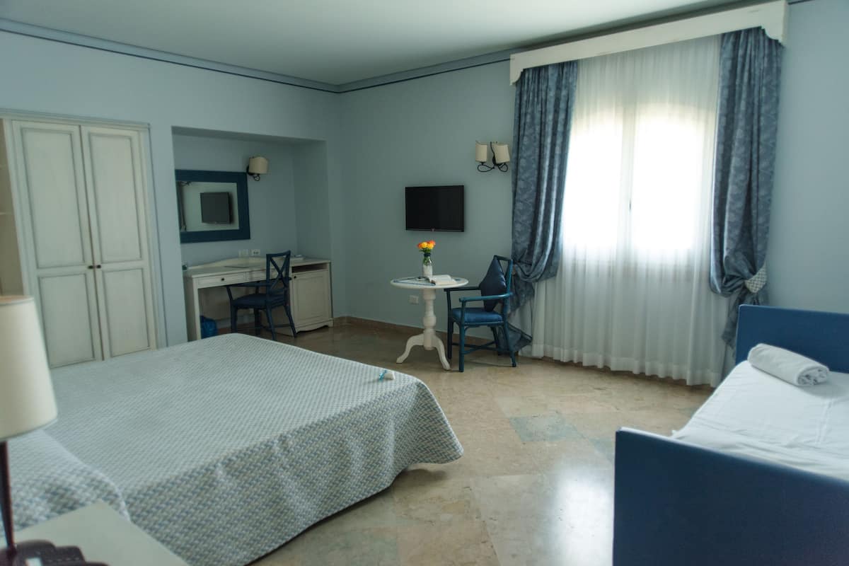 A hotel room with a bed, a desk with a chair, a small table with a vase, and a TV on the wall.