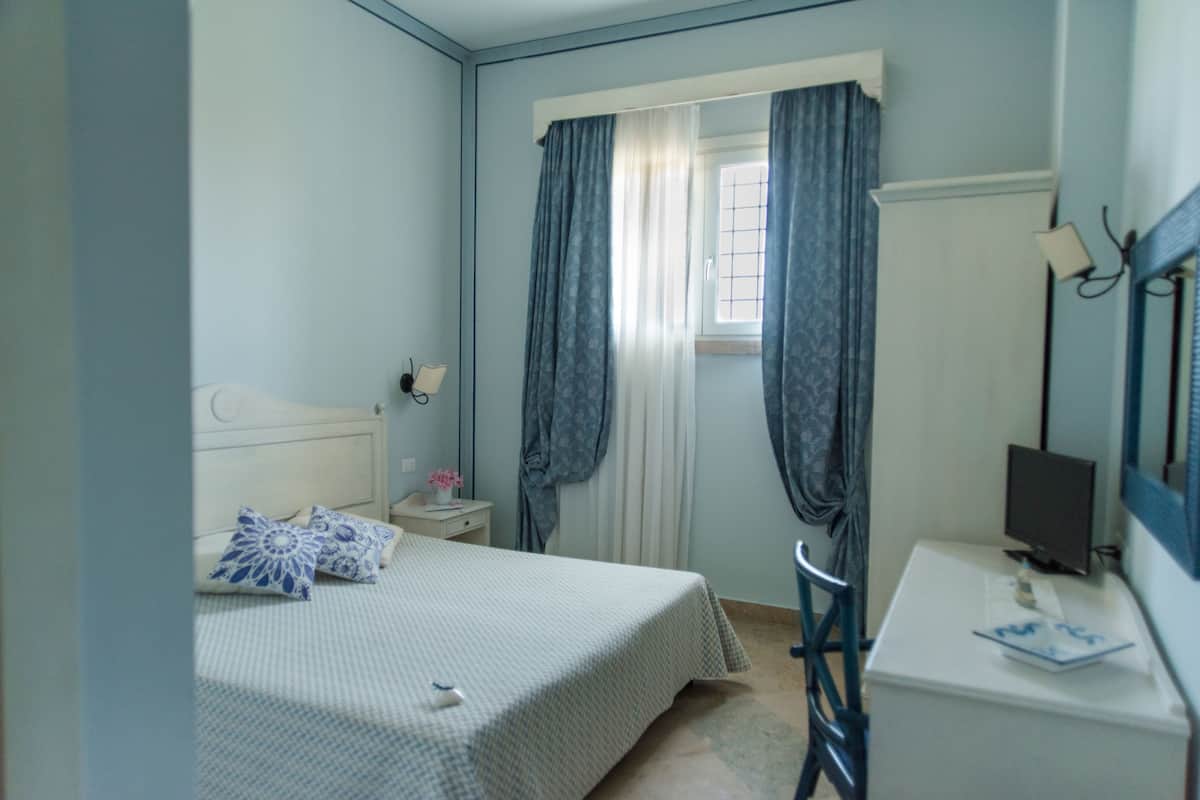 A bedroom with a bed, a desk, a chair, a window with curtains, and a nightstand with a lamp.