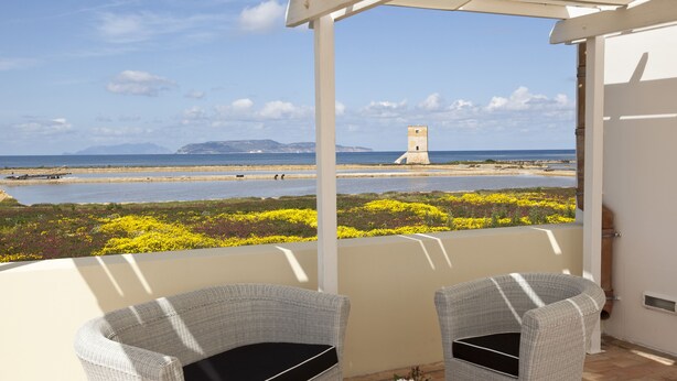 A balcony with wicker chairs, a small table with a flower arrangement, and a view of the sea, a lighthouse, and a distant island. - Relais Antiche Saline (Paceco)