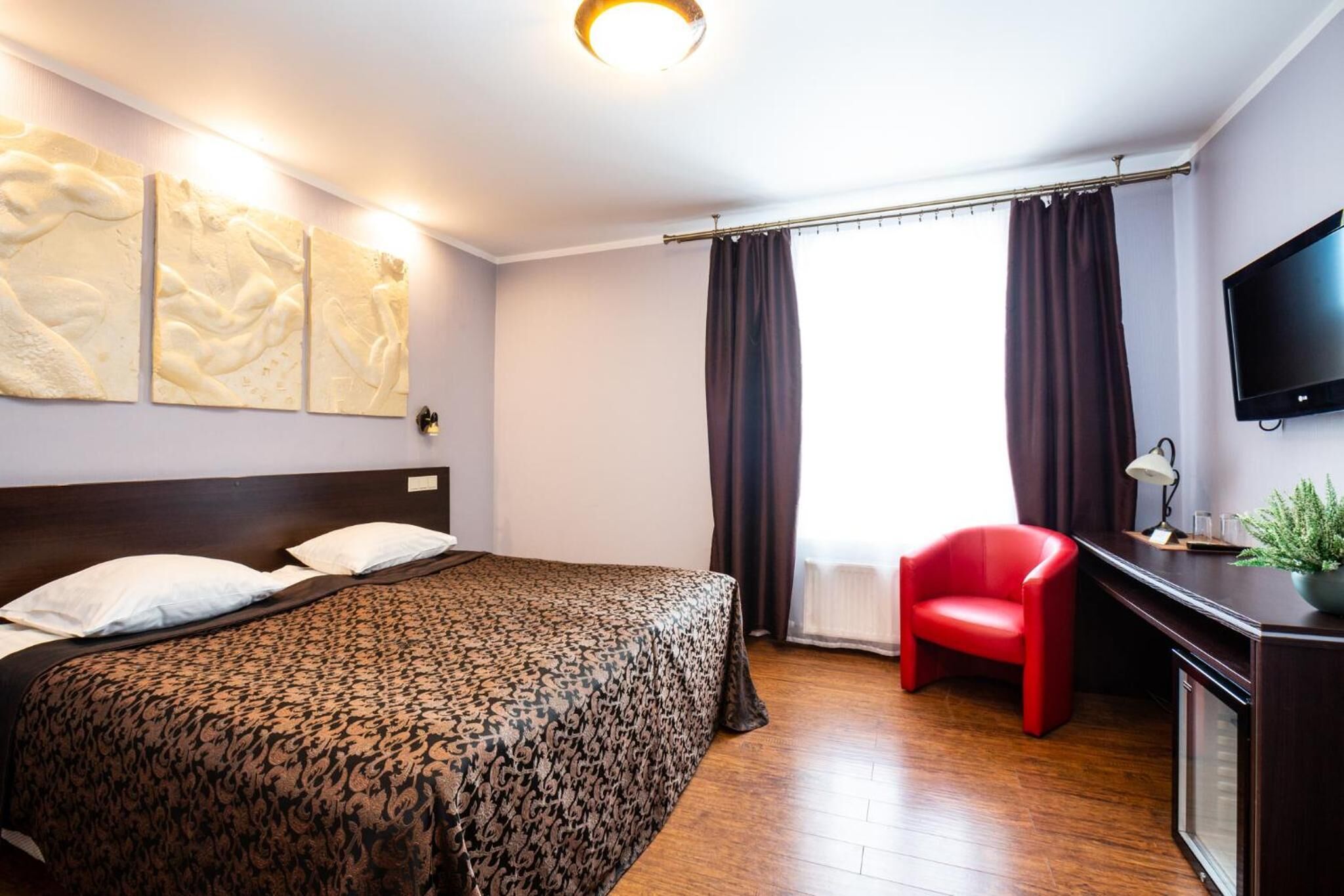 Habitación de hotel con una cama, un escritorio con televisor, una silla roja y obras de arte en la pared.