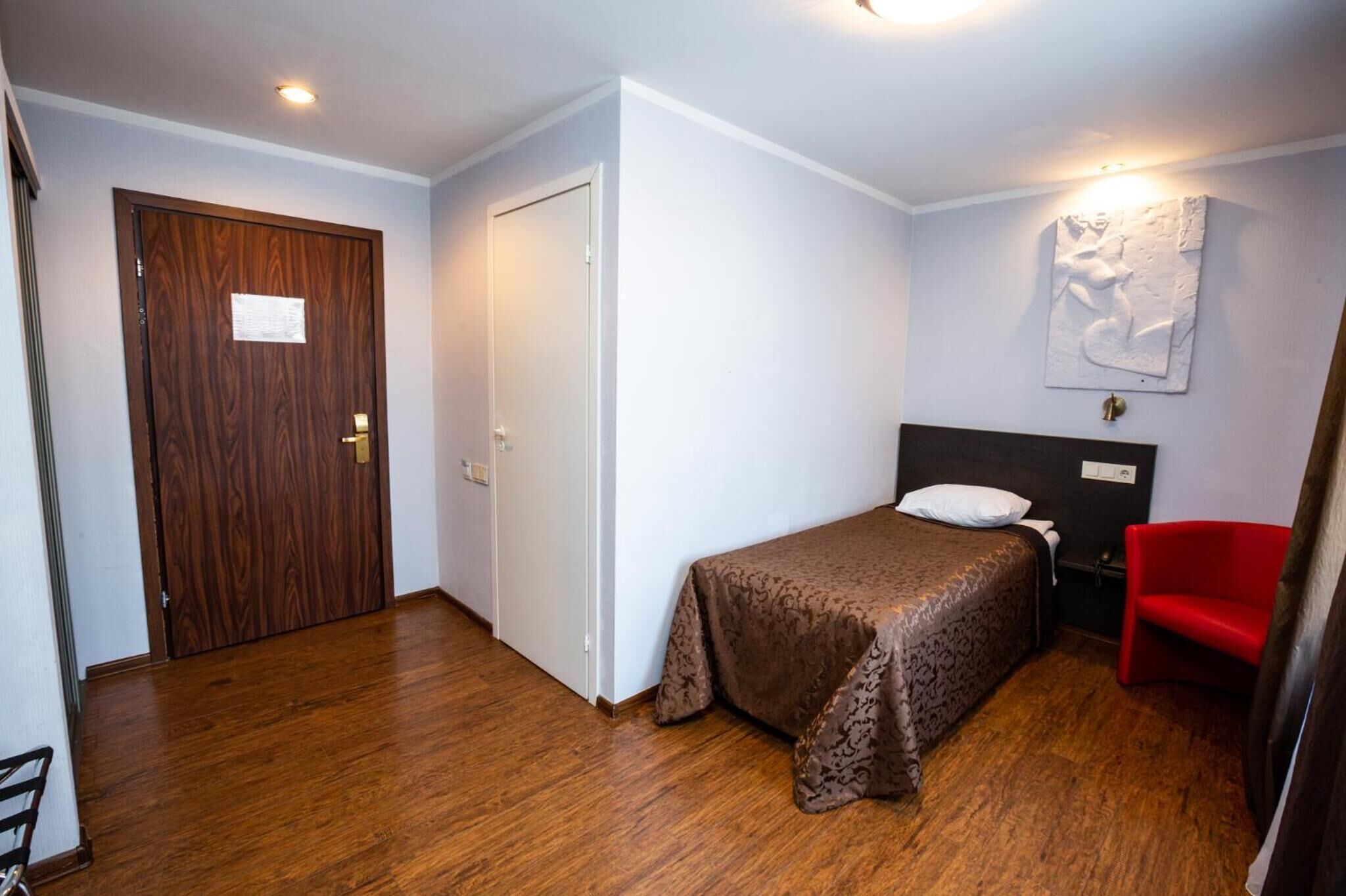 Una habitación pequeña y ordenada con una cama, una silla roja y una puerta de madera.