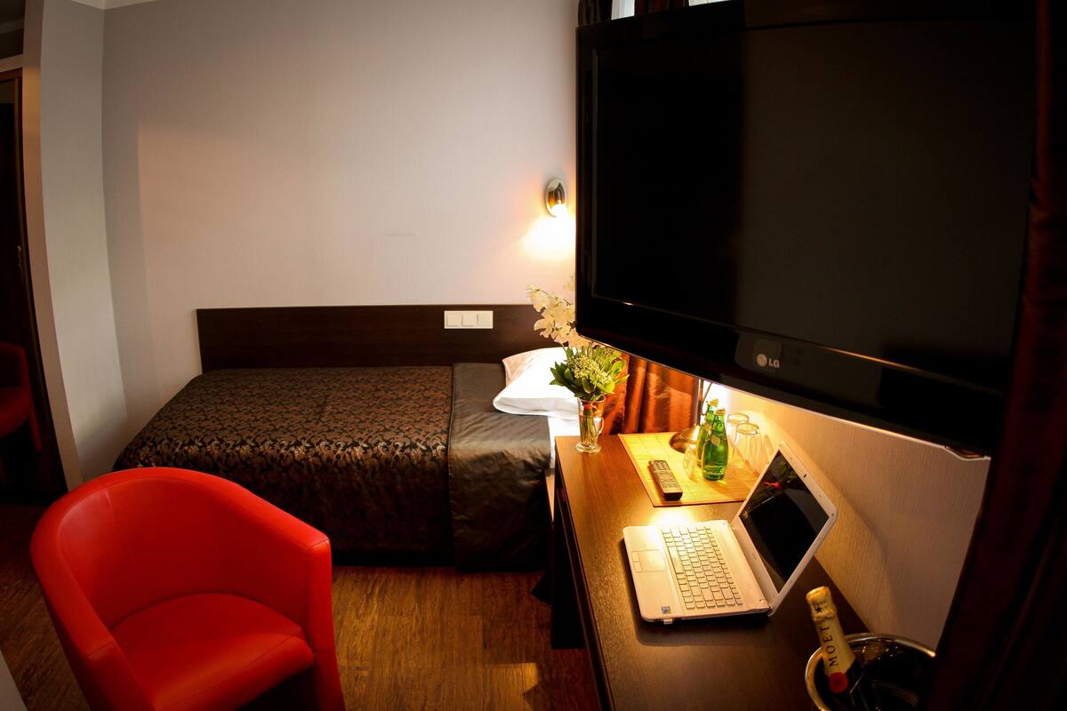 Ein Hotelzimmer mit einem Bett, einem Schreibtisch mit einem Laptop, einem roten Sessel, einem Fernseher und einer Champagnerflasche.