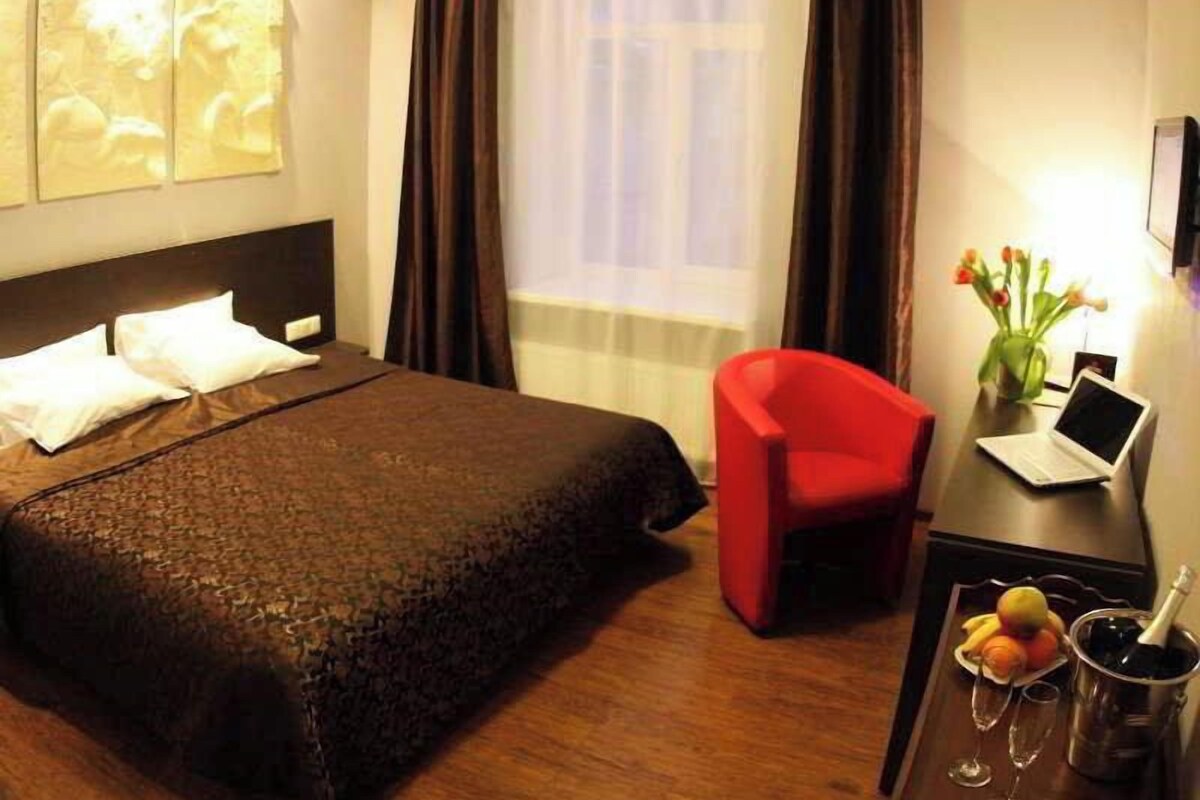 A hotel room with a bed, a red chair, a desk with a laptop, a vase with flowers, and a fruit basket with champagne.