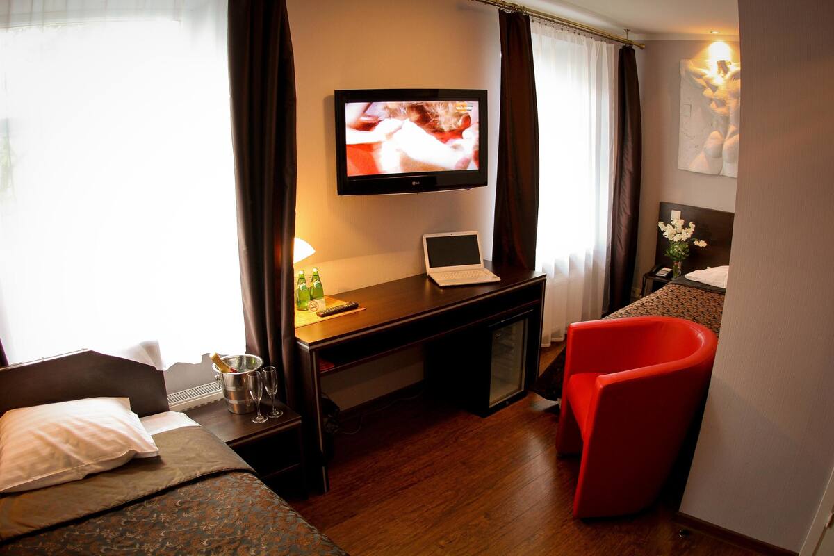 Ein Hotelzimmer mit einem Bett, einem Schreibtisch mit einem Laptop, einem Fernseher, einem roten Sessel und einem kleinen Tisch mit einer Flasche und Gläsern.