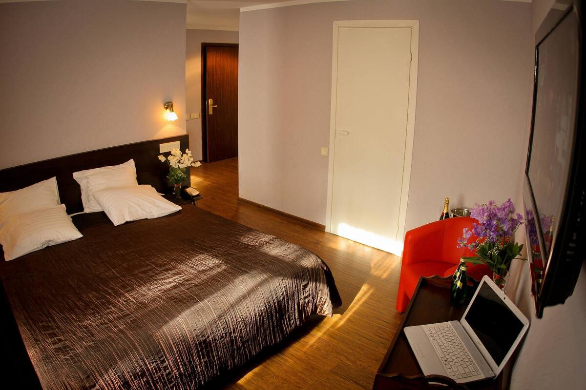 Ein Hotelzimmer mit einem Bett, einem Schreibtisch mit Laptop, einem Sessel, einem Fernseher und einer Tür.
