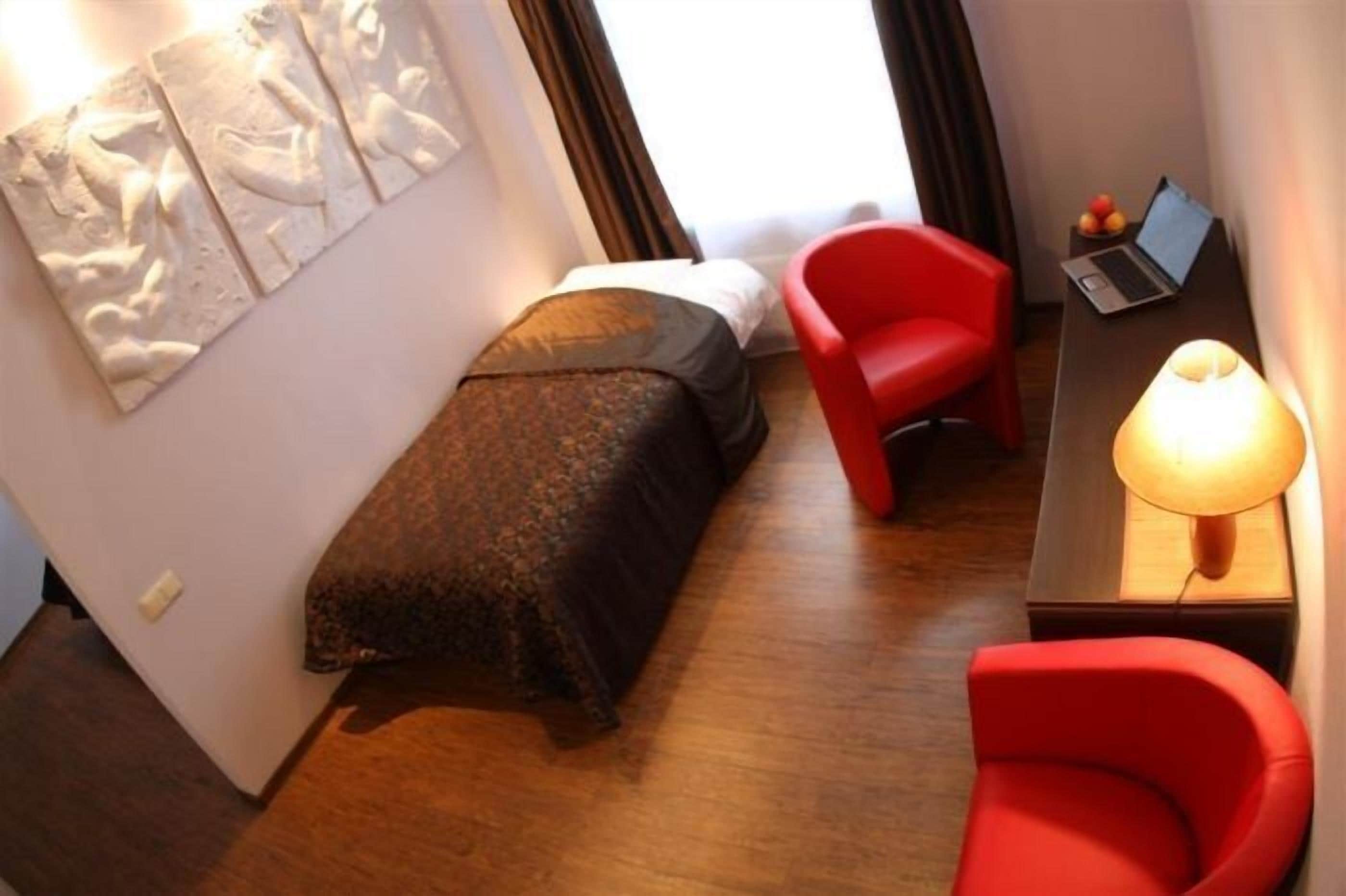 Una habitación pequeña y acogedora con una cama, una silla roja, un escritorio con una computadora portátil y una lámpara, y una obra de arte colgada en la pared.