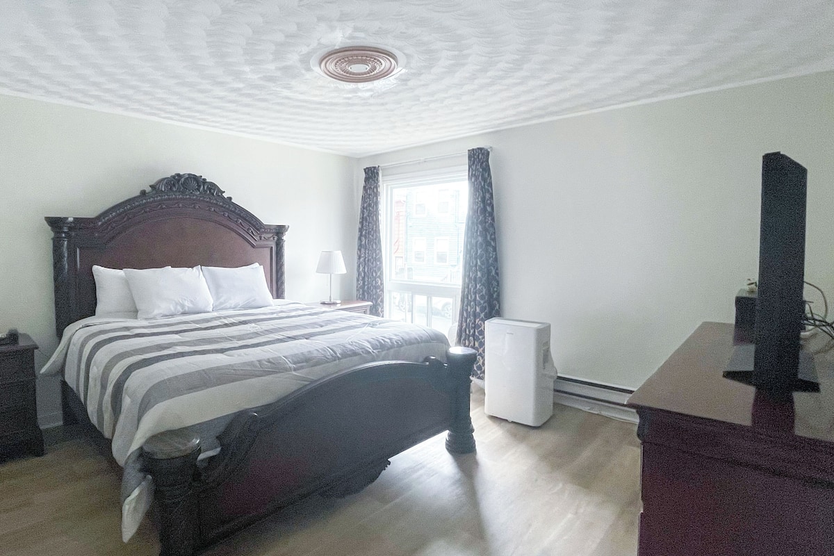 A hotel room with a large bed, a bedside table with a lamp, a dresser, a window with curtains, and a flat-screen TV.