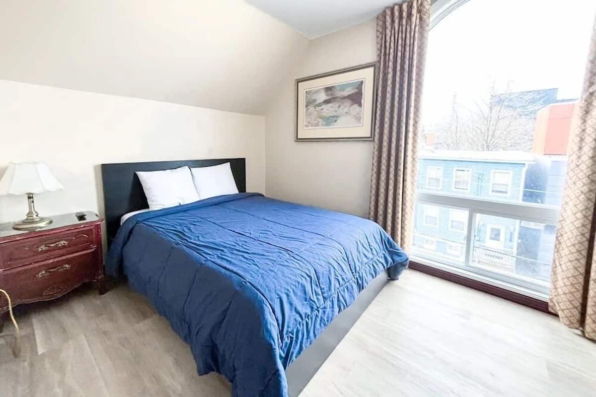 A bedroom with a bed, a nightstand, a lamp, a window with curtains, and a framed picture on the wall.