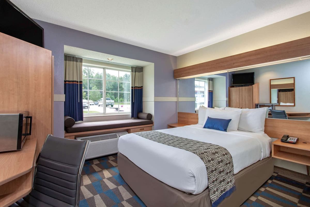 A hotel room with a bed, a desk, a chair, a TV, a window with curtains, and a mirror.
