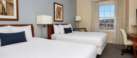 A hotel room with two beds, a desk, a chair, a window with a view, and two wall-mounted lamps.