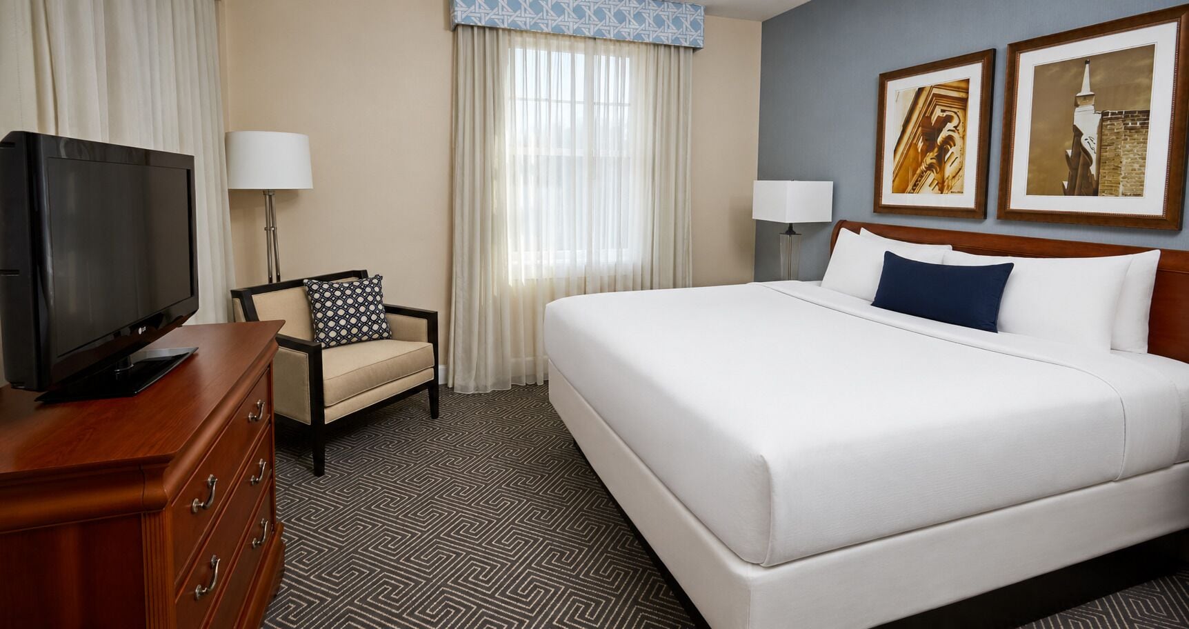 A hotel room with a bed, a television on a wooden dresser, a chair, two lamps, and two framed pictures on the wall.