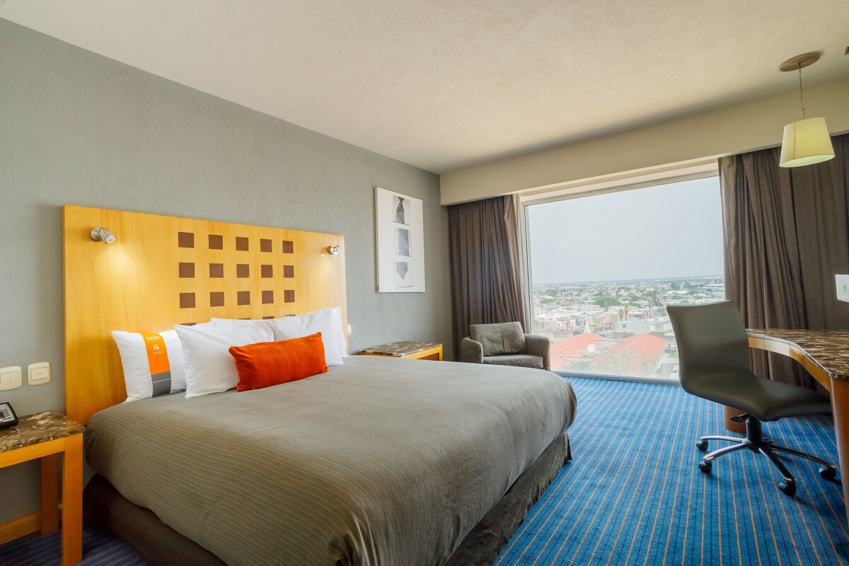 Ein Hotelzimmer mit einem großen Bett, einem Schreibtisch mit Stuhl, Blick auf die Stadt und einem Vorhangfenster.