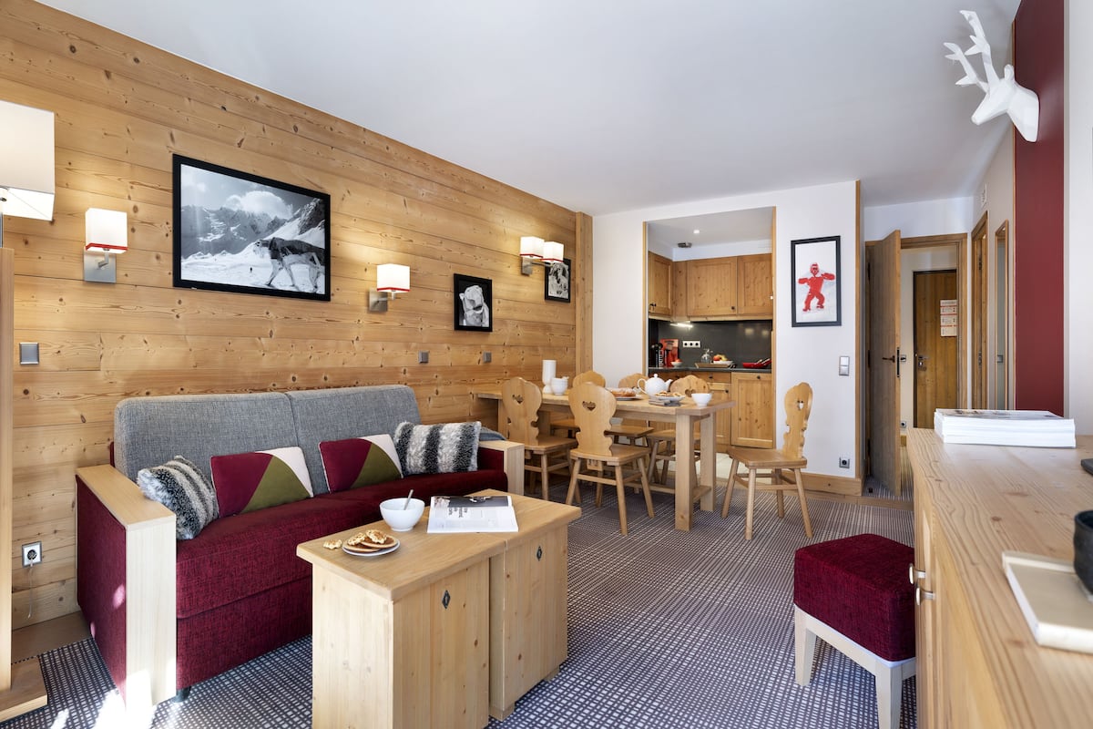 Een gezellige zithoek met een houten wand, een bank, een eettafel en een keuken op de achtergrond.