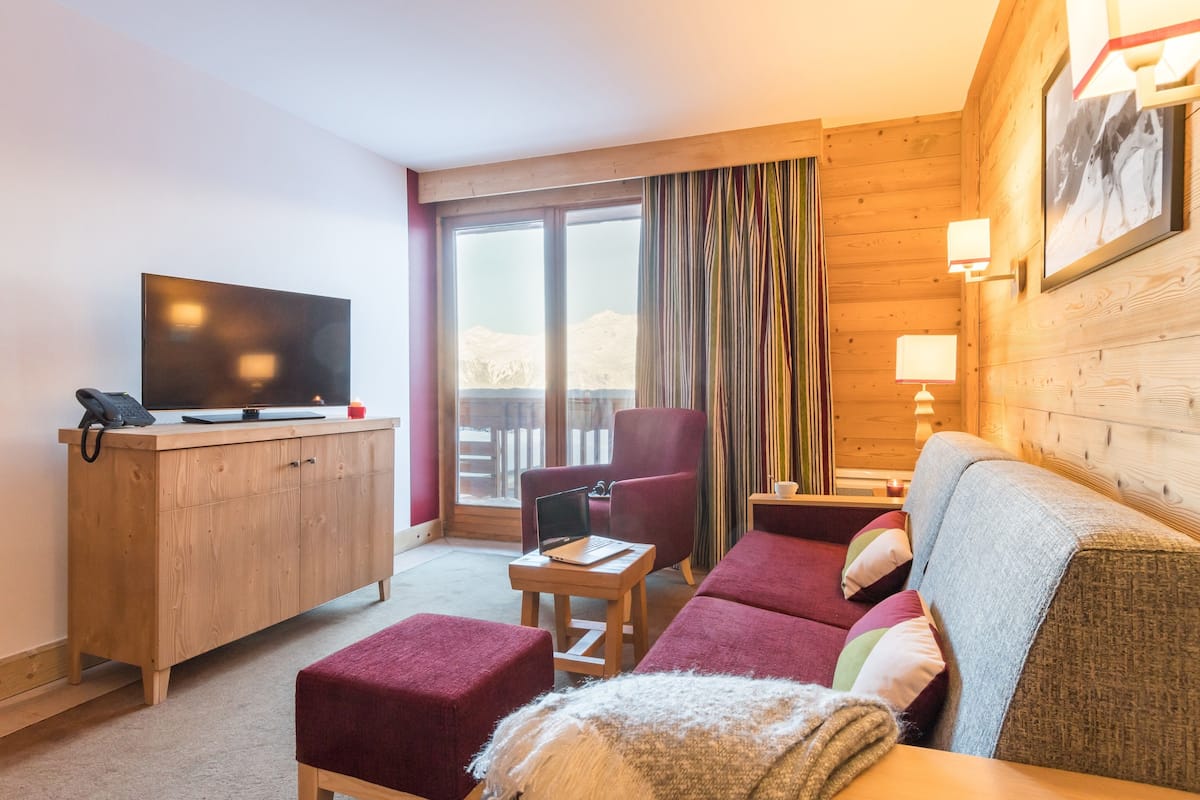 Hotelkamer met houten vloer, een bank, een stoel, een bureau met laptop, een televisie en een raam met gordijnen.