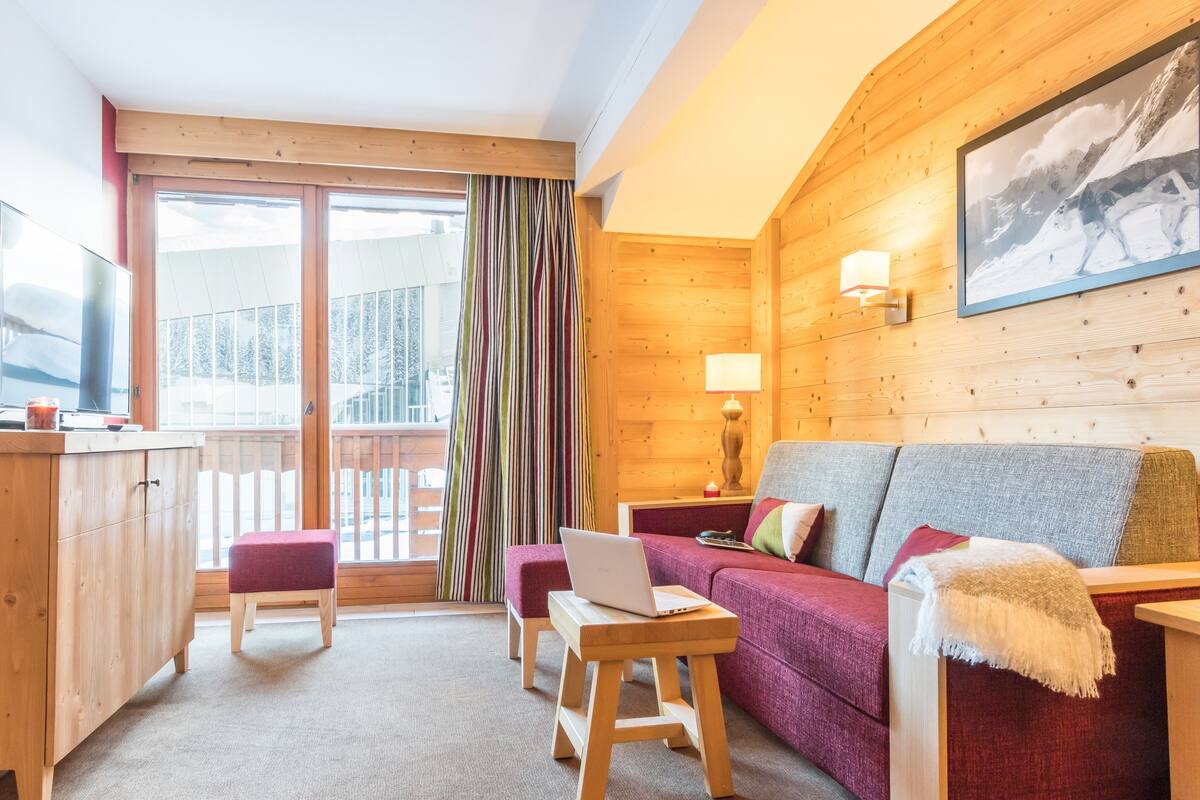 Een knus ingerichte kamer met houten details, een bank, een tafel met een laptop en een televisie op een houten kast.
