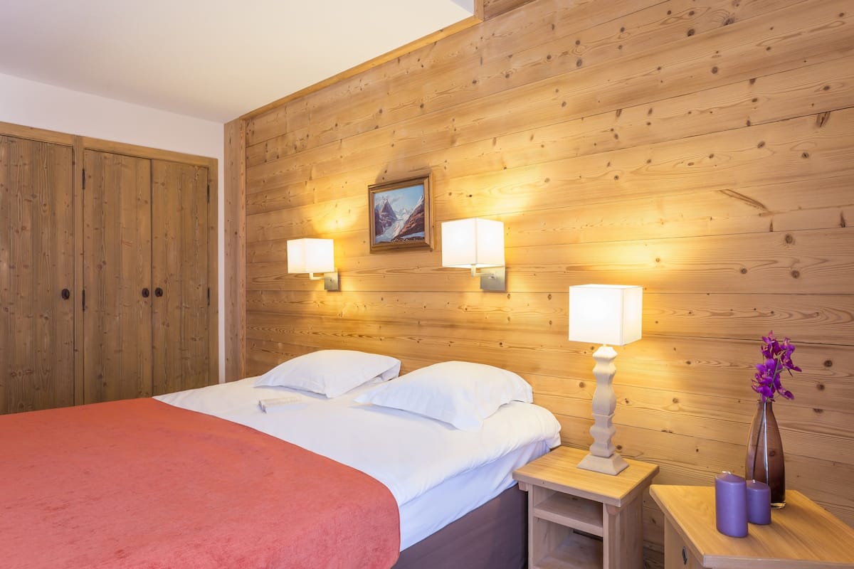 Een kamer met houten lambrisering, een bed, twee bedlampen, een schilderij en een vaas met bloemen.
