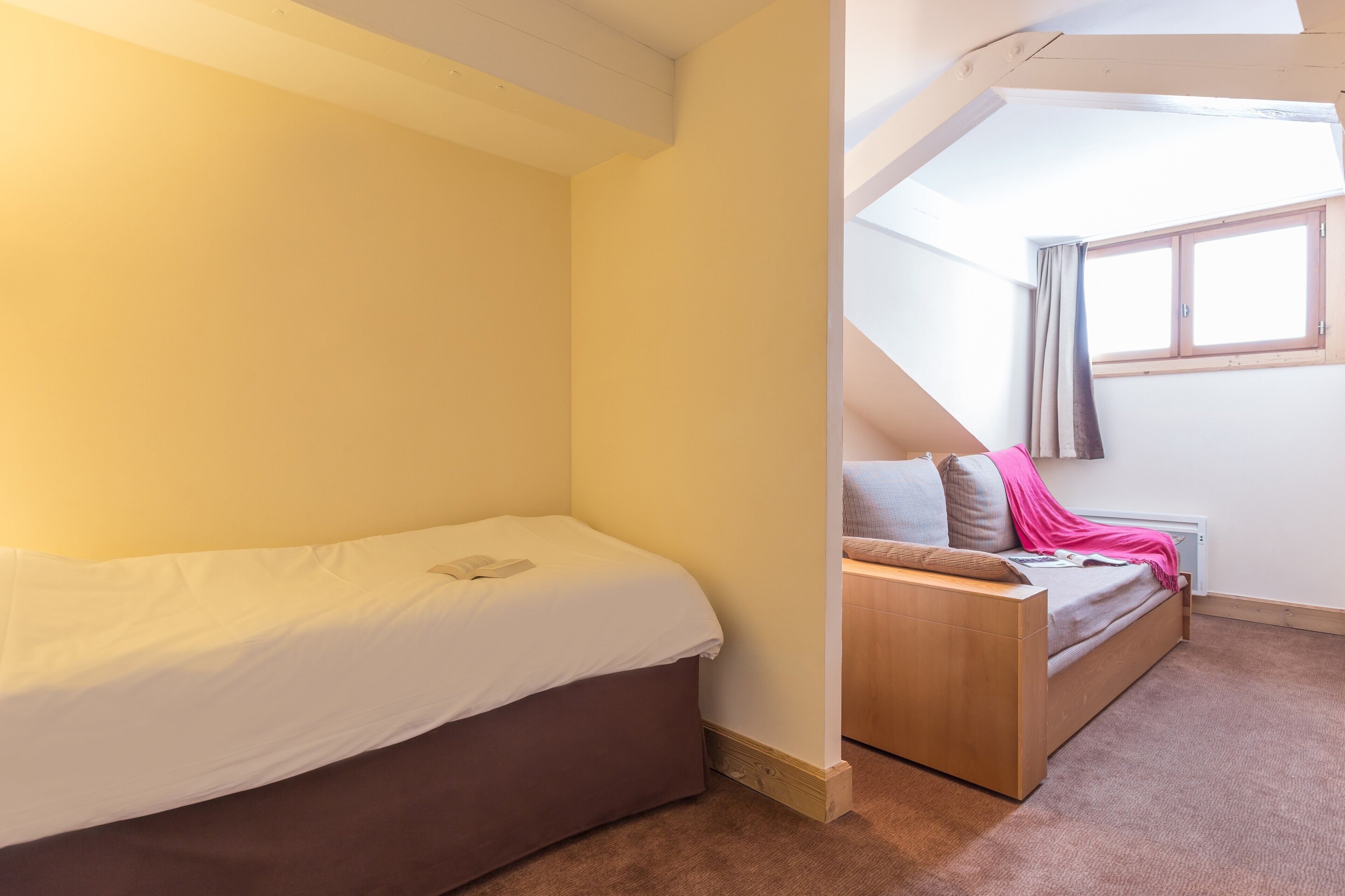 A bedroom with a single bed, a wooden bench, and a window with curtains.
