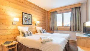 Quarto de hotel com duas camas, paredes de madeira, uma janela com cortinas listradas e uma televisão sobre uma mesa de madeira.