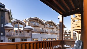 Um deck de madeira com uma cadeira branca e um cobertor de tricô, com vista para um edifício com várias varandas e telhados cobertos de neve.
