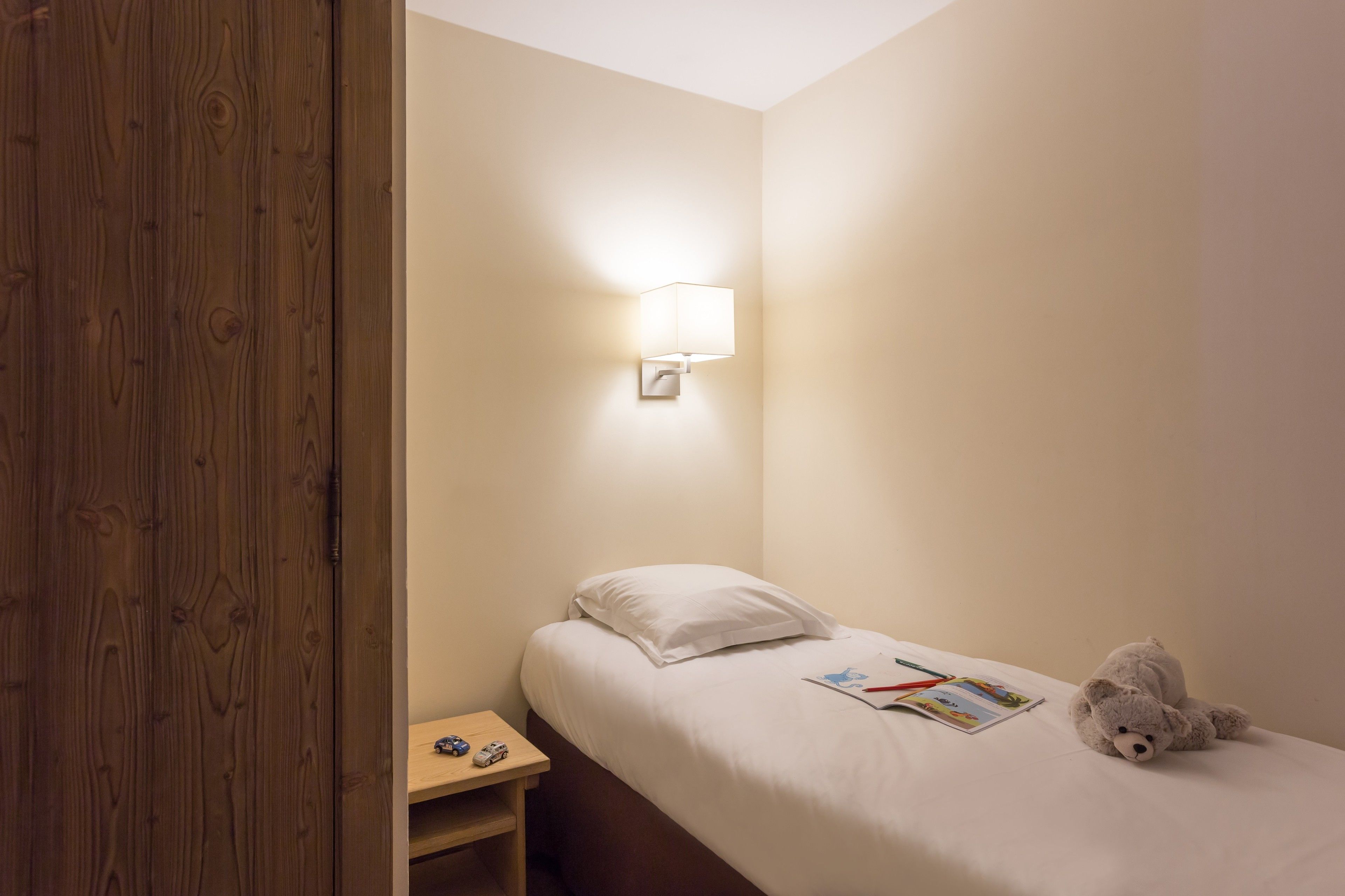 A small, neatly made bedroom with a single bed, a nightstand, a lamp, and a teddy bear.