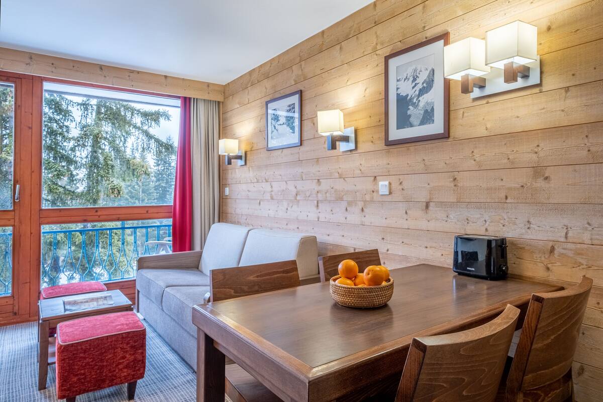 A wooden-paneled room with a dining table, chairs, a sofa, and a window with a view of trees.