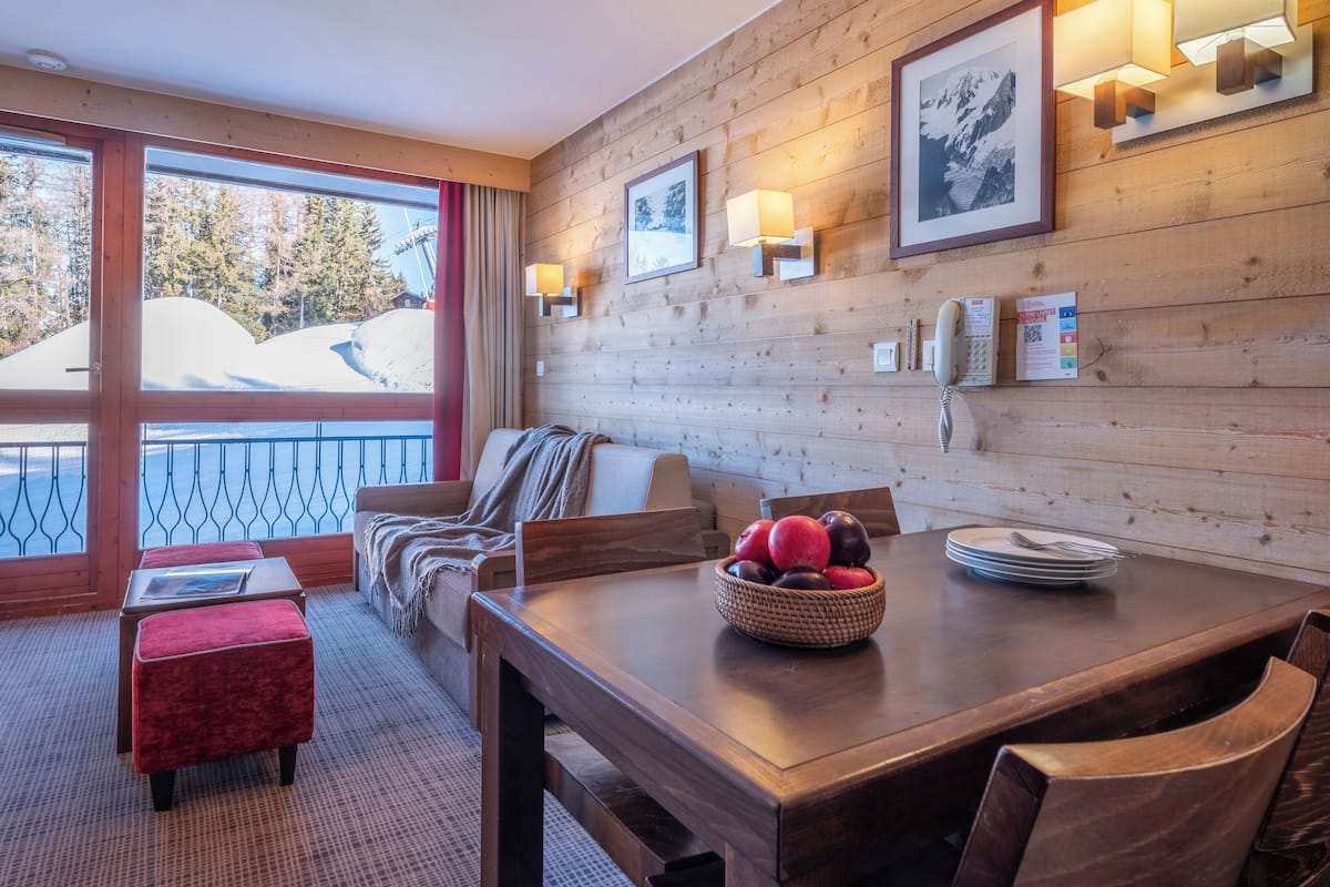 A wooden-paneled room with a dining table, chairs, a red ottoman, and a window with a view of snow-covered mountains.