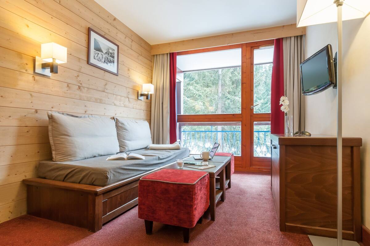 A cozy room with a bed, a small table with a lamp, a TV, and a window with a view of trees.