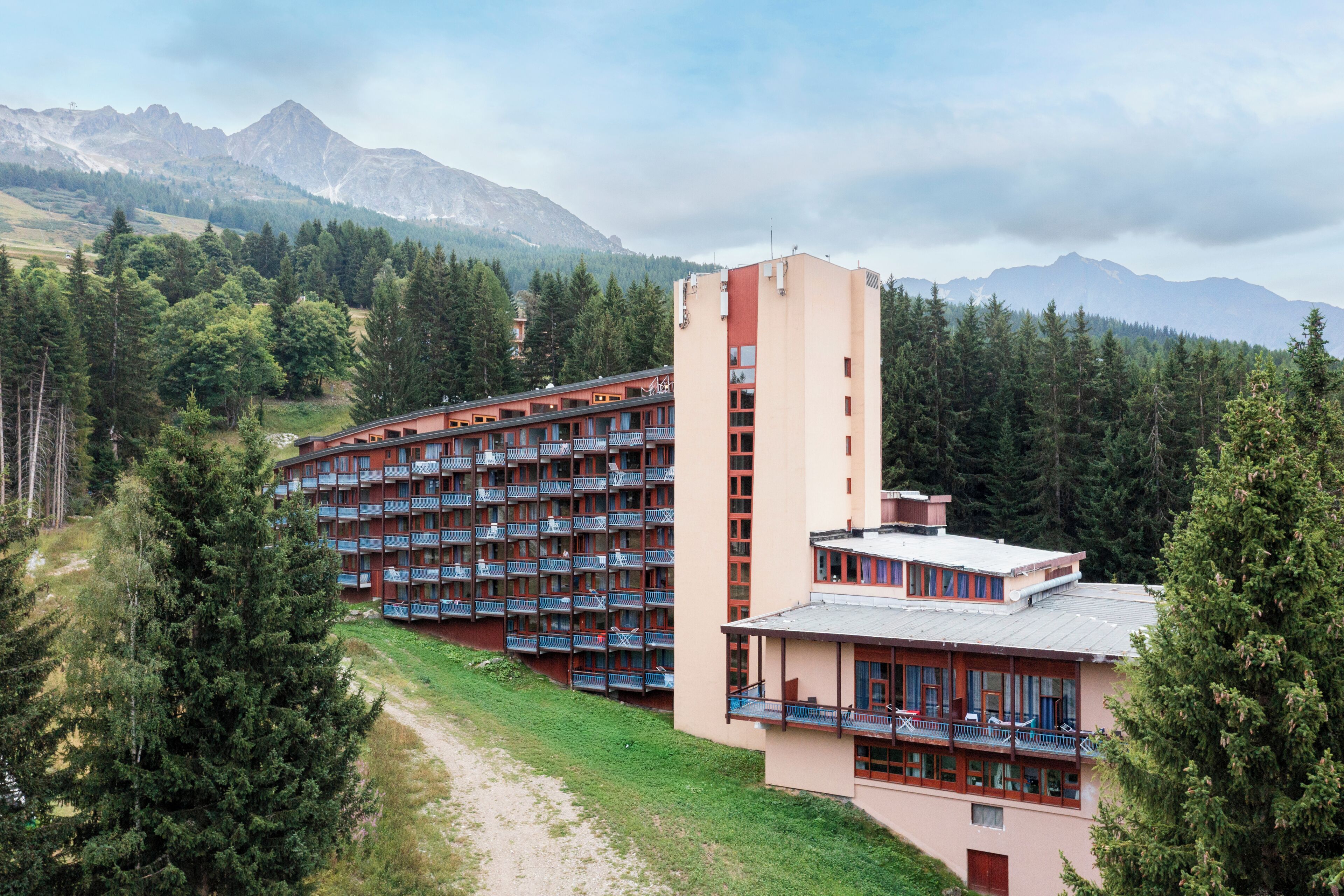 A multi-story hotel with balconies, surrounded by a forest and mountains.