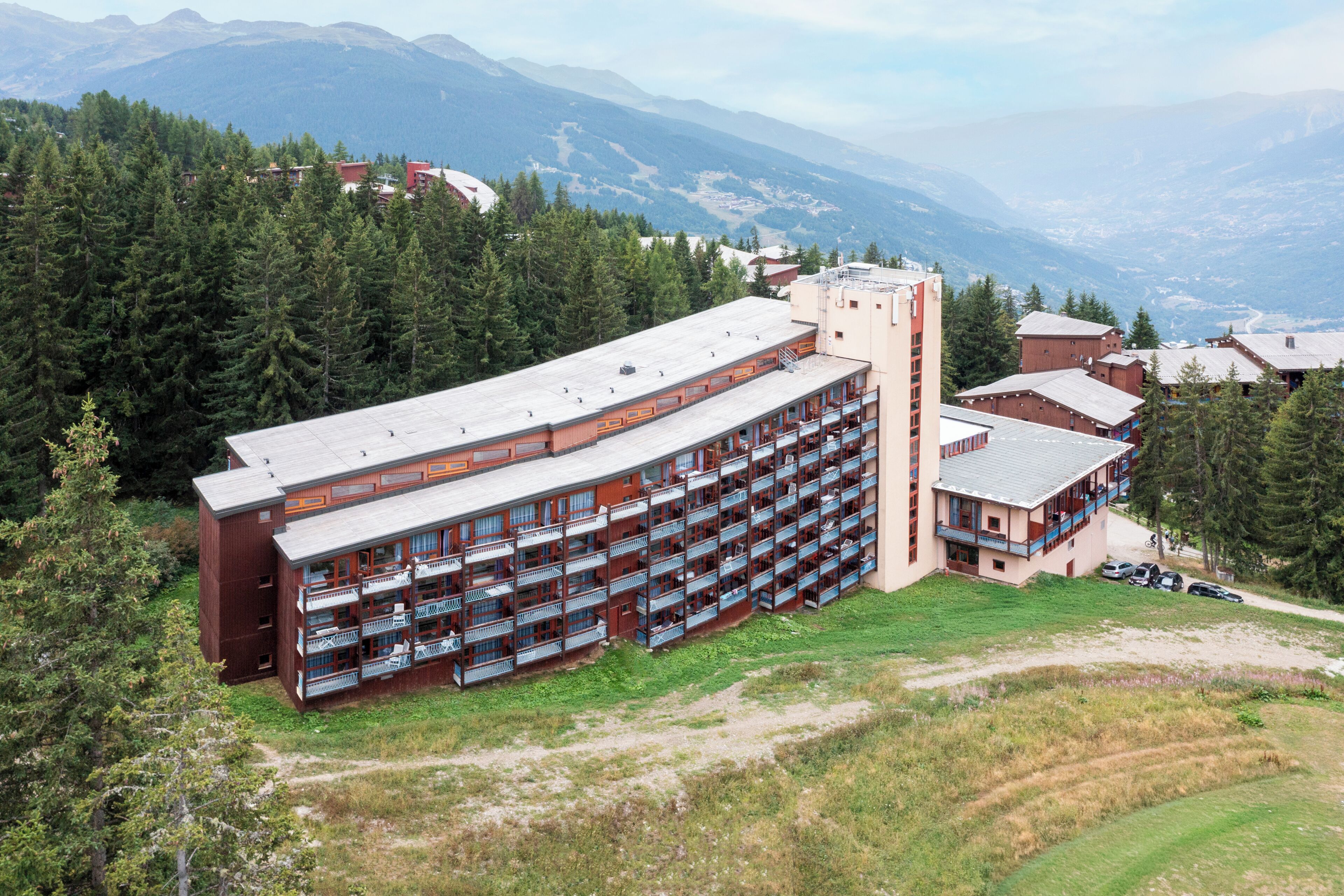 A multi-story hotel with balconies, surrounded by forest and mountains.